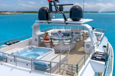 A luxurious yacht with a hot tub in the foreground cruises through clear turquoise waters with a distant, low-lying island in the background.