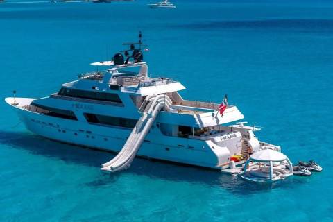 A large white yacht with a slide is anchored in clear turquoise water, with distant boats visible on the horizon.