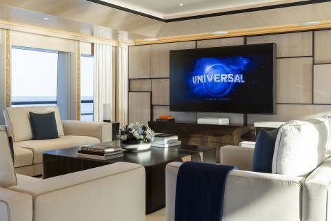 A plush living area with comfortable sofas and a large television displaying the Universal logo is set against a backdrop of ocean views.