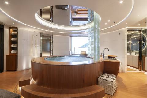 A circular wooden hot tub with a glass column in the center is in the foreground, while a bright, modern spa area with sinks and mirrors is in the background.