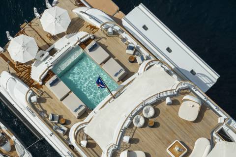 A luxurious yacht with a swimming pool and lounge chairs on its deck floats on the dark blue ocean.