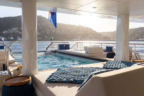 Plush lounge seating and a sparkling pool occupy the foreground, while a sun-drenched coastline with buildings and hills forms the background.