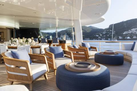 Plush seating and a dining area on a yacht's deck overlook a tranquil bay with a distant village nestled on a hillside.