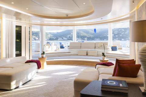 A luxurious yacht interior with plush white sofas and warm accent pillows in the foreground, and a panoramic view of a sun-drenched coastline through large windows in the background.