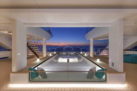 A modern yacht's deck features a sunken seating area in the foreground, with a serene ocean sunset visible in the background.