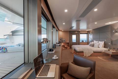 A laptop and coffee sit on a desk in the foreground, while a luxurious bedroom with a large bed and ocean views is in the background.