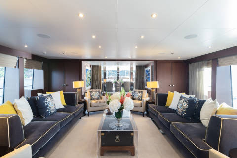 A luxurious yacht lounge with plush sofas and armchairs in the foreground, and a dining area visible through glass doors in the background.