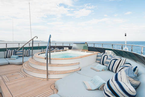 A luxurious yacht's sundeck features a bubbling jacuzzi with a waterfall in the foreground, while the background shows a calm blue ocean under a cloudy sky.