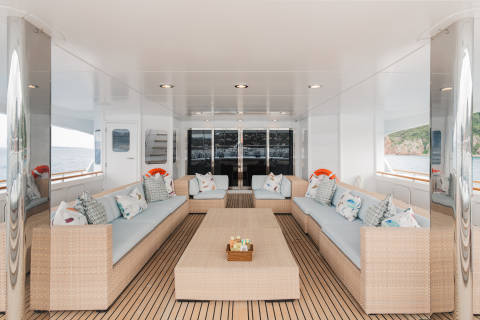 A spacious, light-filled yacht deck with comfortable seating and a coffee table in the foreground, and a scenic ocean view with distant islands in the background.