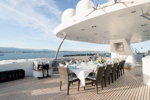 A long dining table set for a meal on a yacht's deck overlooks a calm sea and distant coastline under a bright sky.