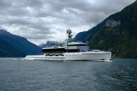 A sleek, modern yacht cruises through calm water with dramatic, cloud-covered mountains rising in the background.