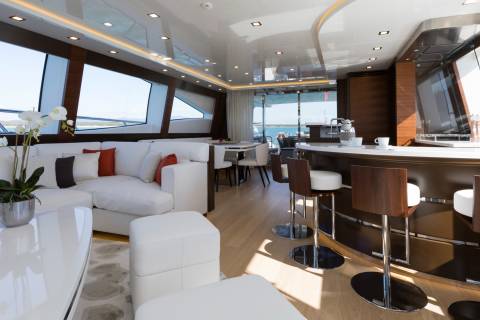 A luxurious yacht interior features a white sectional sofa and ottomans in the foreground, with a polished bar and dining area visible in the background through large windows.