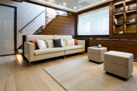 A modern yacht lounge features a cream sofa with colorful throw pillows in the foreground, while the background reveals a wooden staircase and built-in cabinetry.