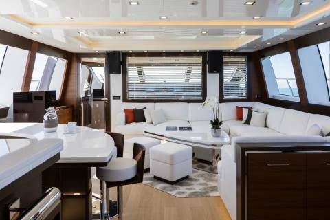 A modern yacht interior features a white bar with stools in the foreground and a spacious white sectional sofa with large windows overlooking the ocean in the background.