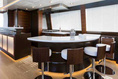 A modern yacht kitchen with a curved island and bar stools in the foreground, and large windows with blinds and a television screen in the background.