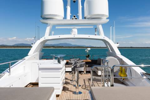A luxurious yacht's deck with comfortable seating and tables is visible in the foreground, while the calm blue ocean stretches to a distant, tree-lined shore under a clear sky in the background.