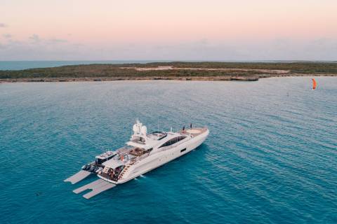 A large white yacht floats on calm turquoise water in the foreground, with a distant, tree-lined island and a kite surfer visible in the pastel-colored sky of the background.