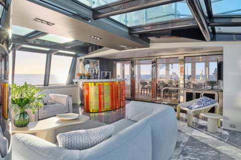 A luxurious yacht interior with plush seating in the foreground and a bar and dining area visible through glass doors in the background, all under a glass ceiling with ocean views.