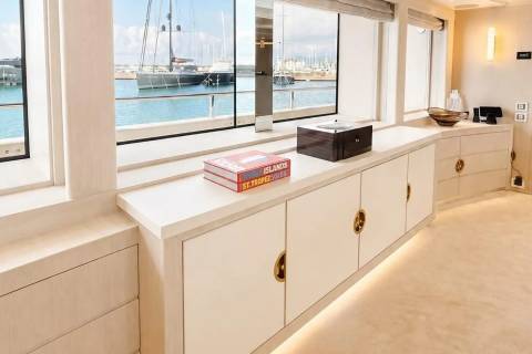 A modern, minimalist interior with books and a dark box on a white cabinet in the foreground, and a large yacht docked in a harbor visible through the window in the background.