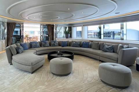 A large, curved sectional sofa with blue and patterned pillows surrounds a dark circular coffee table and two ottomans in the foreground, while large windows offer views of a cityscape and water in the background.
