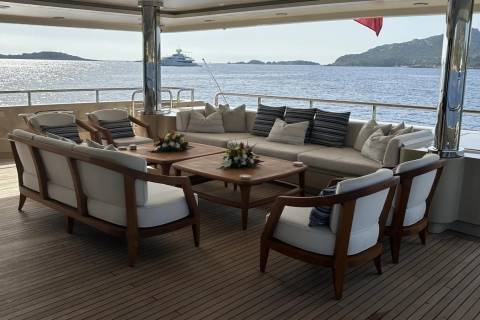 Plush outdoor seating with wooden frames and striped cushions is arranged around coffee tables on a yacht deck, with a calm blue sea and distant islands under a bright sky in the background.