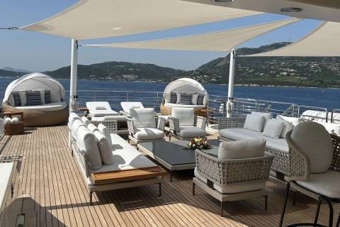 A luxurious yacht deck with comfortable seating and shaded lounging areas is set against a backdrop of a calm blue sea and distant, verdant hills.