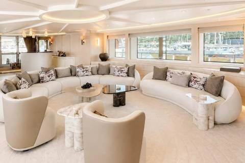 A luxurious, curved cream sofa and modern coffee tables fill the foreground of a bright yacht interior, while large windows in the background reveal a serene view of boats docked in a harbor.