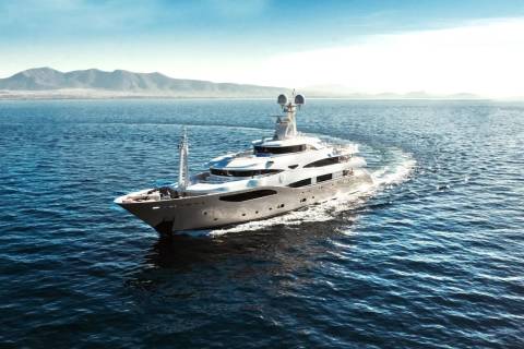 A large, white yacht cruises through calm blue water, leaving a foamy wake, with hazy mountains visible on the horizon under a bright sky.