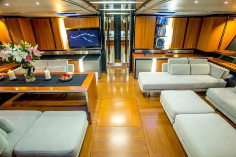 A luxurious yacht interior features a dining table with flowers and candles in the foreground, and plush seating areas and entertainment systems in the background.