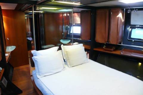 A neatly made bed with white pillows and sheets occupies the foreground, while the background reveals a luxurious cabin with mirrored walls, a sink, and brown leather storage compartments.