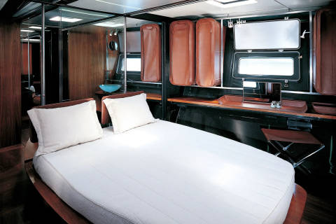 A neatly made bed with white linens dominates the foreground, while the background reveals a compact cabin with mirrored walls, leather storage compartments, and a desk area.