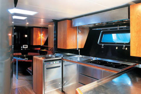 A modern, stainless steel kitchen with wooden cabinets in the foreground is contrasted by a dimly lit dining area with warm lighting in the background.
