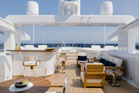 A luxurious yacht deck with a bar and seating area in the foreground overlooks a calm blue ocean under a clear sky.