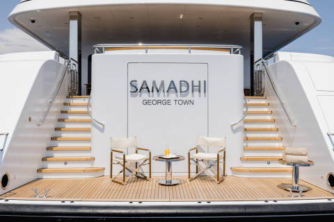 Two white director's chairs with a small table between them sit on the wooden deck of a large white yacht, with the name "SAMADHI" visible on the stern.