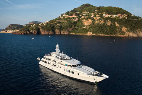 A large white yacht floats on calm blue water in the foreground, with a lush green, hilly coastline dotted with houses in the background.
