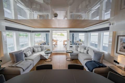 Plush sectional sofas and small tables fill the foreground of a luxurious yacht's lounge, while large windows reveal a serene waterfront view in the background.
