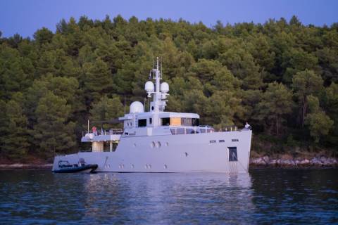 A large, white yacht is anchored in calm water in front of a dense, green forest.