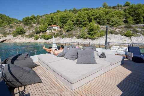 A person relaxes on a large sunbed on a yacht's deck, with a lush, tree-covered hillside and a small building in the background.