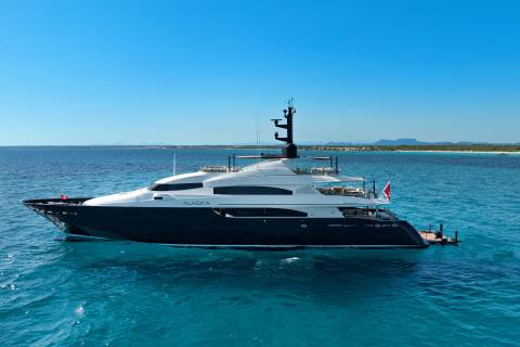 A large, dark-hulled yacht named "ALASKA" floats on turquoise water in the foreground, with a distant, hazy coastline under a clear blue sky in the background.