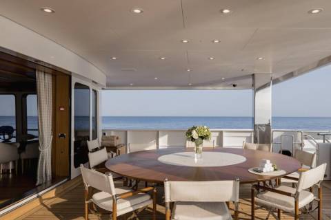 A circular dining table with chairs on a wooden deck is set against a backdrop of the calm blue ocean.