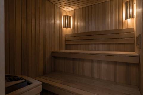 Warm wooden benches and walls fill the foreground of a dimly lit sauna, with two glowing wall sconces illuminating the background.