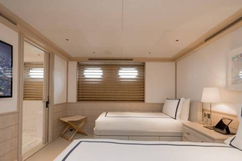 Two neatly made twin beds with navy trim are in the foreground, while the background reveals a bathroom with a porthole window and a framed artwork on the wall.