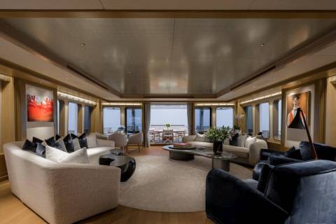 Plush sofas and modern art adorn the spacious, well-lit living area of a yacht, with large windows offering a glimpse of the ocean and distant horizon.