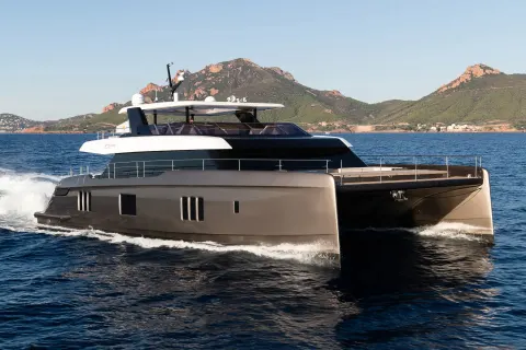 A sleek, modern catamaran yacht cruises through calm blue water with a rugged, mountainous coastline in the background.