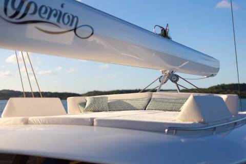 A white sailboat mast with the name "Hyperion" curves over a plush seating area on a yacht's deck, with a calm blue sea and distant green hills under a clear sky in the background.