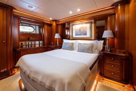 A luxurious yacht cabin features a plush bed with decorative pillows in the foreground, while the background reveals polished wood paneling and a framed nautical painting.