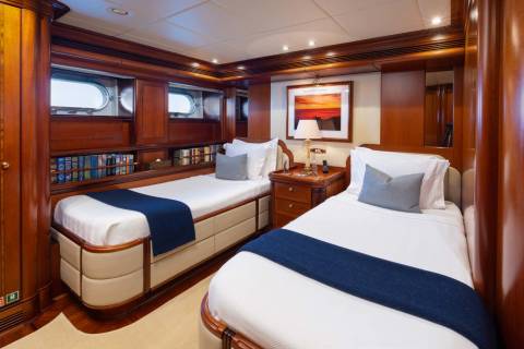 Two neatly made twin beds with blue runners and white linens are in the foreground, while the background features wood-paneled walls, porthole windows, and a framed artwork above a nightstand.