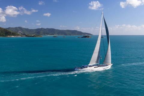 A white sailboat with full sails glides across turquoise water, leaving a foamy wake, with a lush green coastline and distant islands under a bright blue sky in the background.