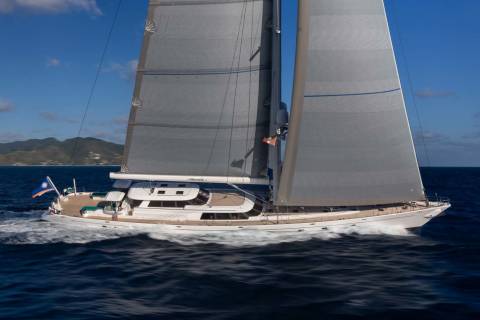 A large white sailboat with full sails glides through dark blue water, with green, hilly islands visible in the hazy background under a bright blue sky.