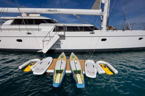 Four paddleboards and two yellow jet skis are docked on a wooden platform in the foreground, with a large white yacht docked behind them.